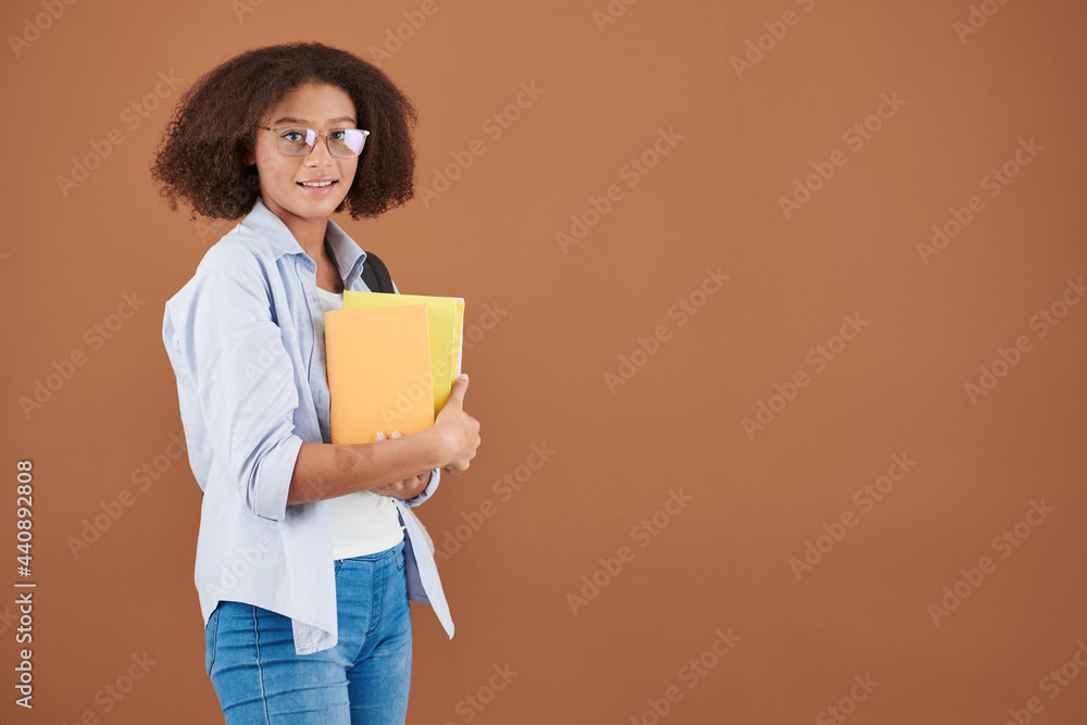 Obraz premium Portrait of smiling mixed-race teenage girl in eyeglasses standing with yellow books against brown background