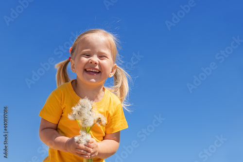 A little charming girl with dandelions is laughing against the blue sky. Summer sunny child.