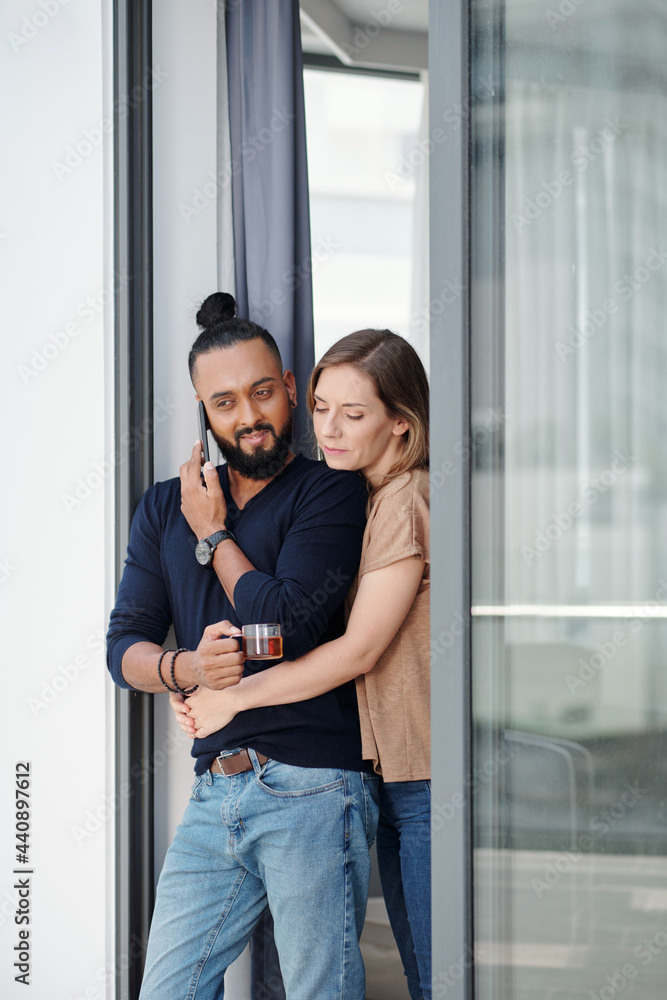Naklejka premium Pretty young woman hugging her husband drinking tea and talking on phone with coworker or employee