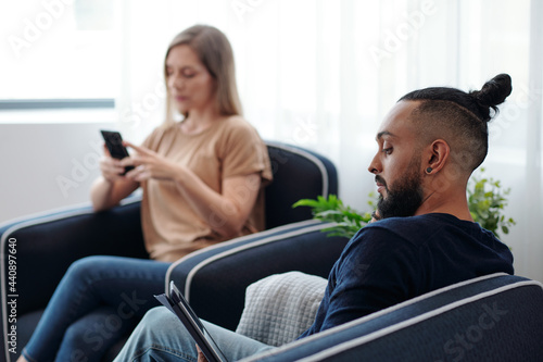 Concentrated bearded man reading e-book, his wife checking text messages on smartphone in background