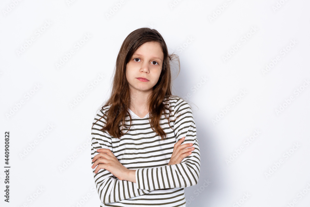 A sad dark-haired teenage girl stands with her arms crossed. On a light background. Portrait. Place for your text. A sad dark-haired teenage girl stands with her arms crossed. On a light background. Portrait. Place for your text.