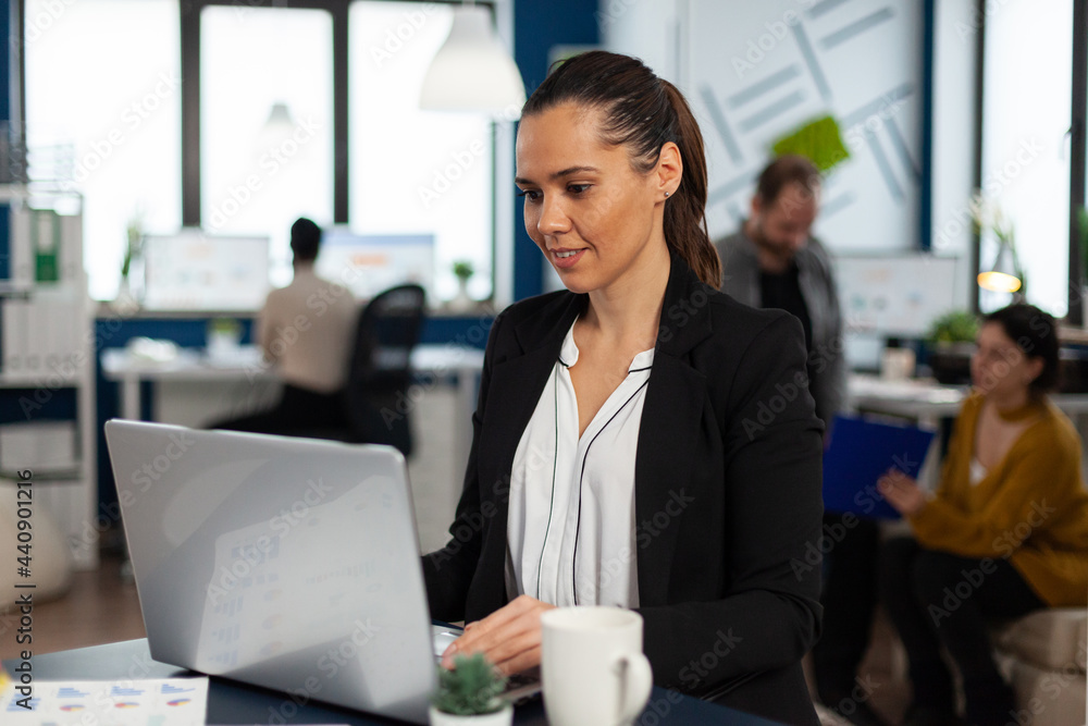 Enthusiastic woman manager typing on laptop, browsing on internet while sitting at desk smiling receiving good news. Multiethnic coworkers talking about startup financial company in modern office Enthusiastic woman manager typing on laptop, browsing on internet while sitting at desk smiling receiving good news. Multiethnic coworkers talking about startup financial company in modern office