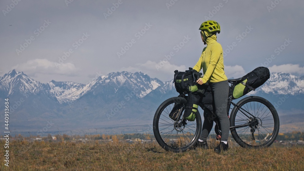 Fototapeta premium The woman travel on mixed terrain cycle touring with bikepacking. The traveler journey with bicycle bags. Sport tourism bikepacking, bike, sportswear in green black colors. Mountain snow capped.