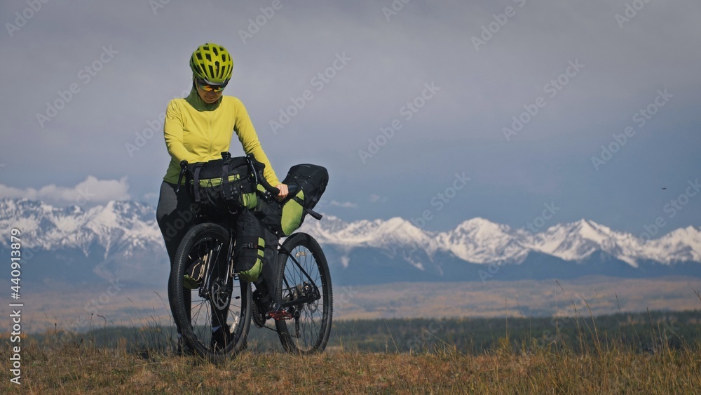 The woman travel on mixed terrain cycle touring with bikepacking. The traveler journey with bicycle bags. Sport tourism bikepacking, bike, sportswear in green black colors. Mountain snow capped. The woman travel on mixed terrain cycle touring with bikepacking. The traveler journey with bicycle bags. Sport tourism bikepacking, bike, sportswear in green black colors. Mountain snow capped.