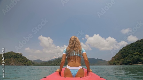 Sexy young woman in blue swimsuit swings feet in water on pink canoe on sea against hills backside view. Traveling to tropical countries. Attractive sportive girl is sailing on kayak in ocean. © ivandanru