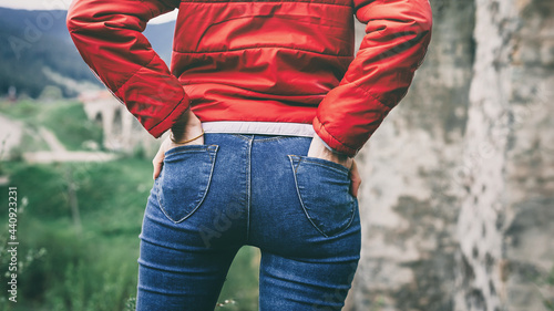 Young woman in stylish jeans. A girl in a red jacket and blue jeans holds her hands in her back pockets. Outdoors.