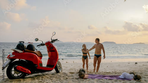 Scooter road trip. Lovely couple on red motorbike in white clothes on sand beach. Just married people kiss hugs walking near the tropical palm trees, sea. Wedding honeymoon by ocean. Motorcycle rent. © ivandanru
