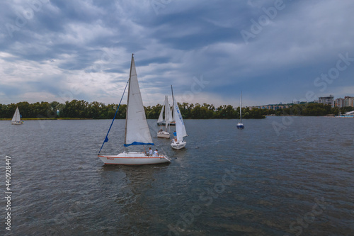 Regatta of sailing yachts with white sails on the lake in the city park. Aerial view of a sailboat in windy conditions. Summer sport yachting. Swimming in cloudy weather.