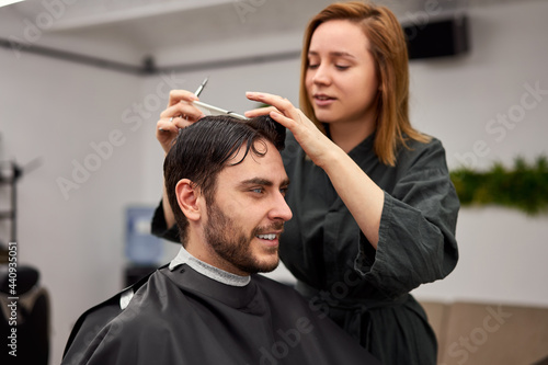 Handsome blue eyed man sitting in barber shop. Hairstylist Hairdresser Woman cutting his hair. Female barber.