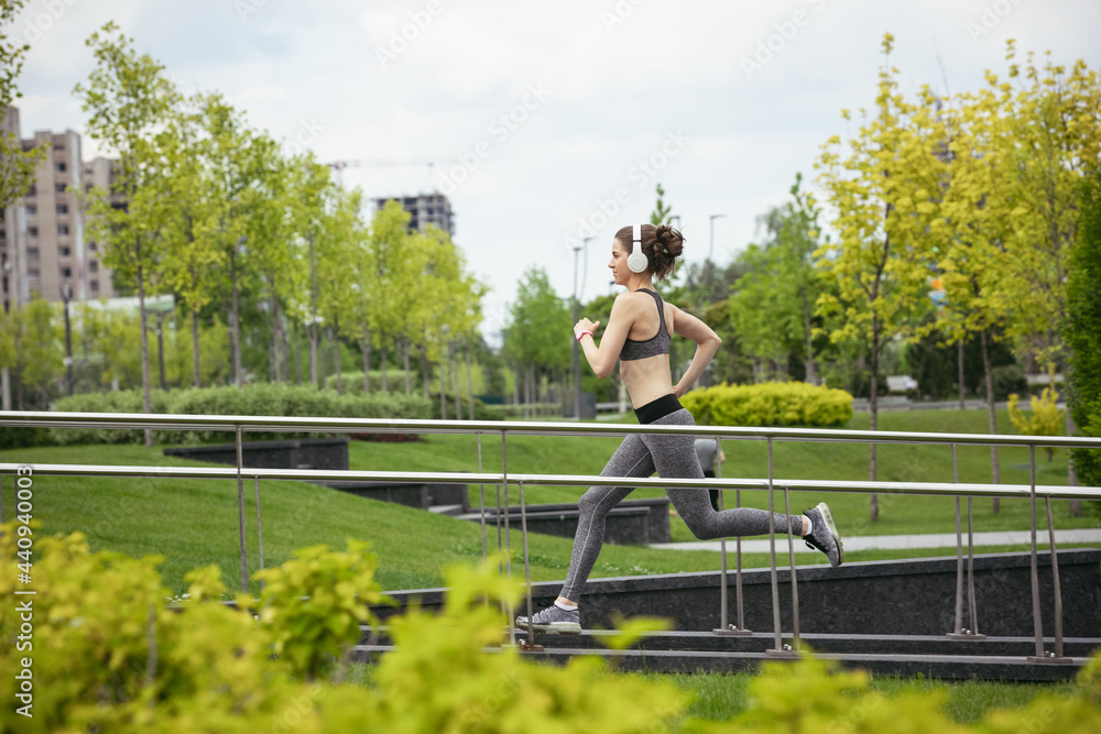 Fototapeta premium Young female runner, athlete is jogging in the city street in spring sunshine. Beautiful caucasian woman training, listening to music. Concept of sport, healthy lifestyle, movement, activity.