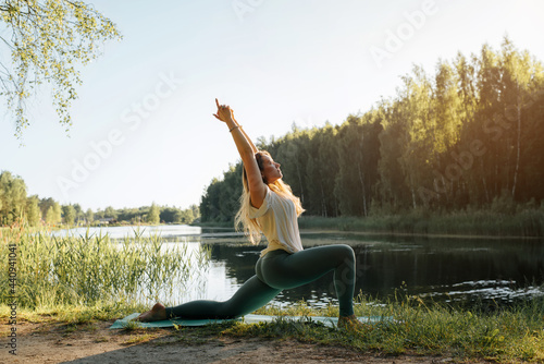 Yoga in nature. Young woman practicing yoga on lake at sunset, summer. Fitness model standing on sports mat in anjaneyasana pose, performing yoga asana, outdoors. Active healthy lifestyle concept