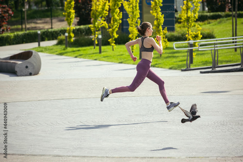 Young female runner, athlete is jogging in the city street in spring sunshine. Beautiful caucasian woman training, listening to music. Concept of sport, healthy lifestyle, movement, activity. Young female runner, athlete is jogging in the city street in spring sunshine. Beautiful caucasian woman training, listening to music. Concept of sport, healthy lifestyle, movement, activity.