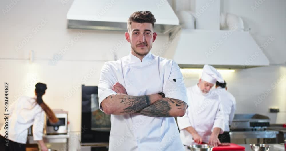 Camera approaching handsome happy young Caucasian male chef standing in restaurant kitchen at work looking at camera in good mood. Portrait of man chef. People working on background Camera approaching handsome happy young Caucasian male chef standing in restaurant kitchen at work looking at camera in good mood. Portrait of man chef. People working on background