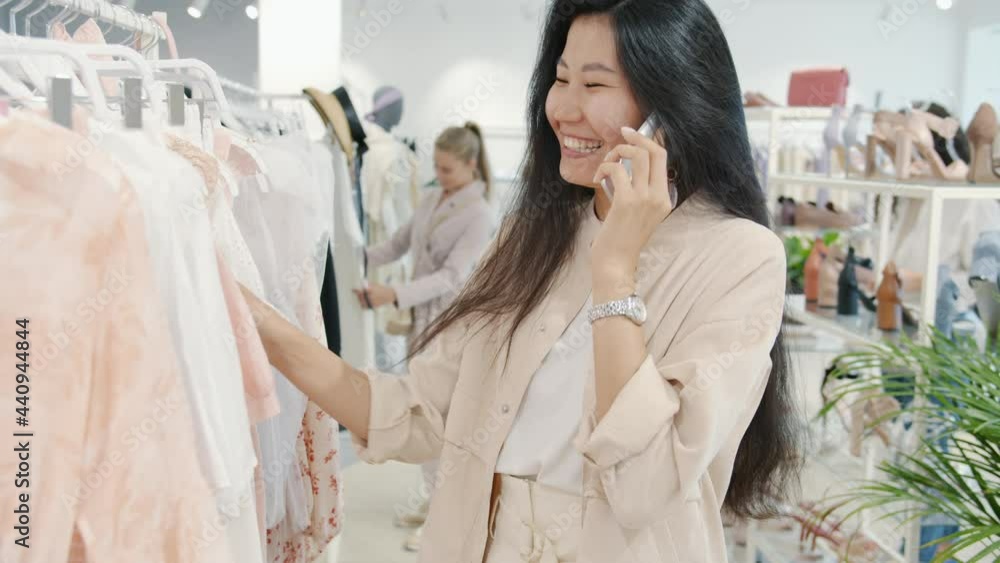 Attractive Asian woman is chatting on mobile phone smiling and looking at clothes in fashion shop enjoying modern communication and shopping Attractive Asian woman is chatting on mobile phone smiling and looking at clothes in fashion shop enjoying modern communication and shopping