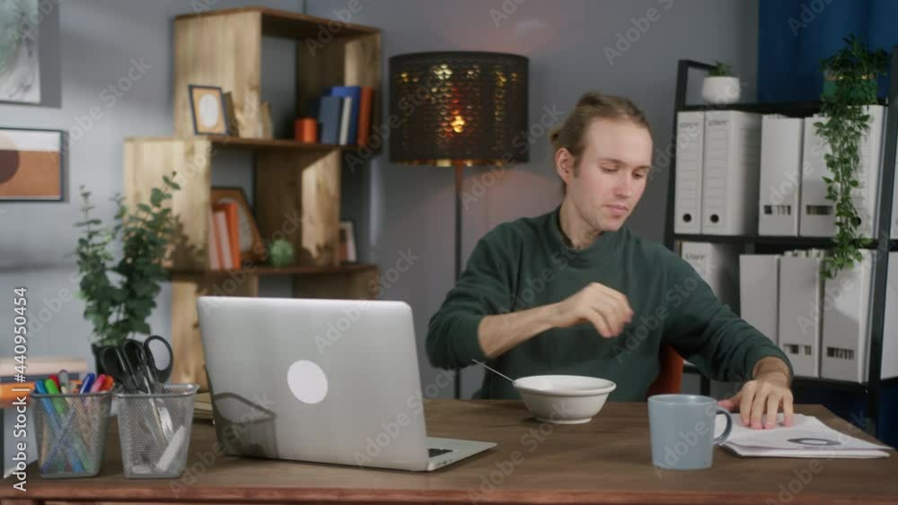 Young man is getting down to eat during break at workplace in office. Freelancer opening laptop, eating muesli, cereal, having breakfast in early morning, lunch, watching video on computer. Young man is getting down to eat during break at workplace in office. Freelancer opening laptop, eating muesli, cereal, having breakfast in early morning, lunch, watching video on computer.
