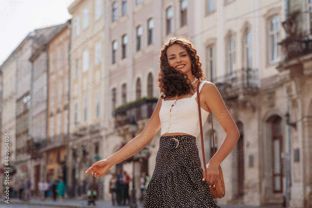 Happy smiling fashionable curly woman wearing trendy summer outfit posing in street of European city. Fashion, lifestyle conception. Copy, empty space for text