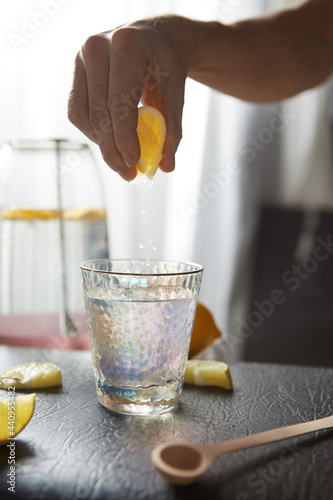 Cropped shot of female hand squeezing lemon juice into clear drinking glass with golden rim. Conical cocktail glass with hammered texture is covered rainbow tinting. Photo is made in the kitchen.  photo