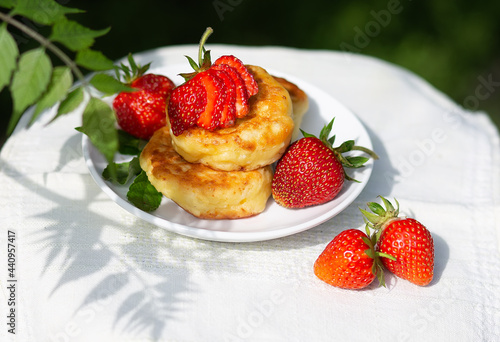 Beautiful summer breakfast - cheesecakes pancakes with fresh strawberries on a white plate. Tea in a tnka porcelain cup  sun glare and shadows. Linen tablecloth  rustic style  provence