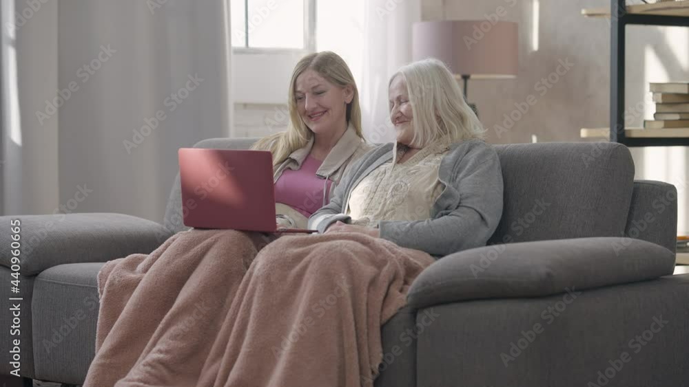 Wide shot portrait of two laughing senior Caucasian women surfing social media on laptop talking. Happy cheerful beautiful old ladies enjoying meeting in living room indoors gossiping. Friendship Wide shot portrait of two laughing senior Caucasian women surfing social media on laptop talking. Happy cheerful beautiful old ladies enjoying meeting in living room indoors gossiping. Friendship