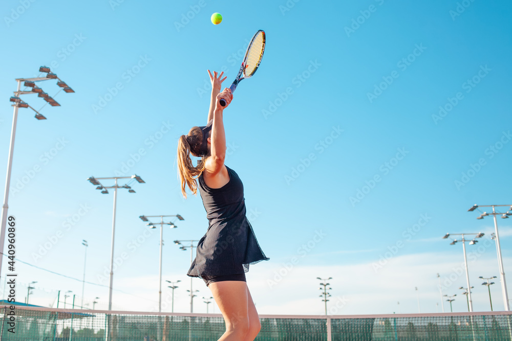 A beautiful woman plays tennis on the court. Sporting a black suit dress. Blue sky, empty space for text. The girl is giving the ball. Exercise, active lifestyle, concept. portrait. close up