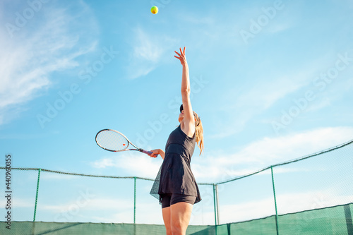 A beautiful woman plays tennis on the court. Sporting a black suit dress. Blue sky, empty space for text. The girl is giving the ball. Exercise, active lifestyle, concept. portrait. outdoor © Valeriia