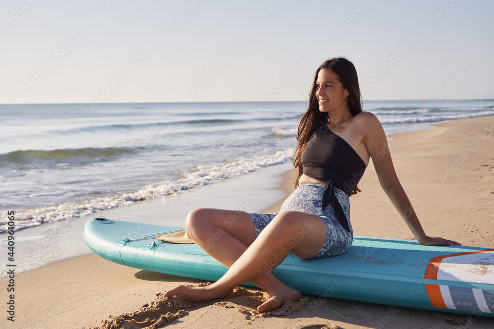 Happy young woman watching the sunrise sitting on a paddle board on the beach