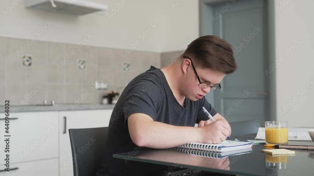 A focused man wearing glasses with down syndrome is writing something sitting at the table at home A focused man wearing glasses with down syndrome is writing something sitting at the table at home