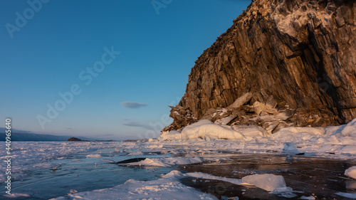 A whimsical rock, devoid of vegetation, against the backdrop of a blue sky. Snow on sheer granite slopes and on the ice of a frozen lake. Reflection on the surface. Evening golden hour. Baikal