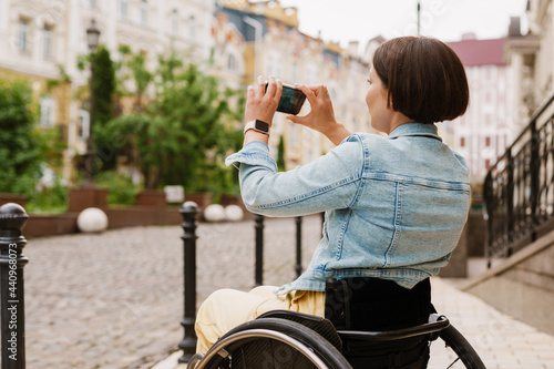 Brunette woman taking photo on mobile phone while sitting in wheelchair