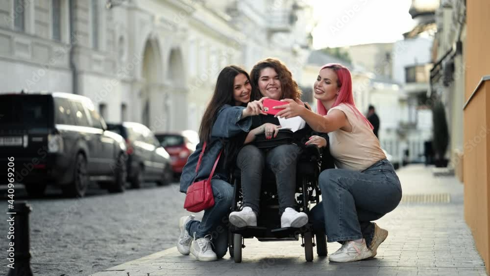 A cheerful disabled woman is sitting on the wheelchair while spending time with her friends and taking selfie photo on the city street A cheerful disabled woman is sitting on the wheelchair while spending time with her friends and taking selfie photo on the city street
