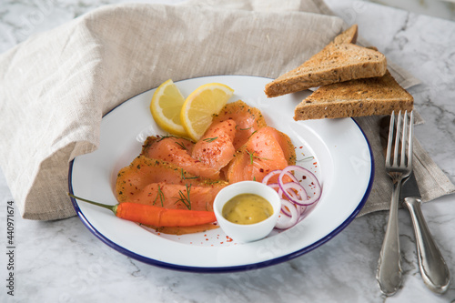 Asia Art Gravlax Graved Lachs, ein roh gebeizter Fisch mit Zitronengras und Chili auf Emaille Teller mit Hovmästarsås Senf Soße, Toast, Zwiebel Ringe, Dill, Zitrone und Leinen Tuch auf hell Marmor Hin photo