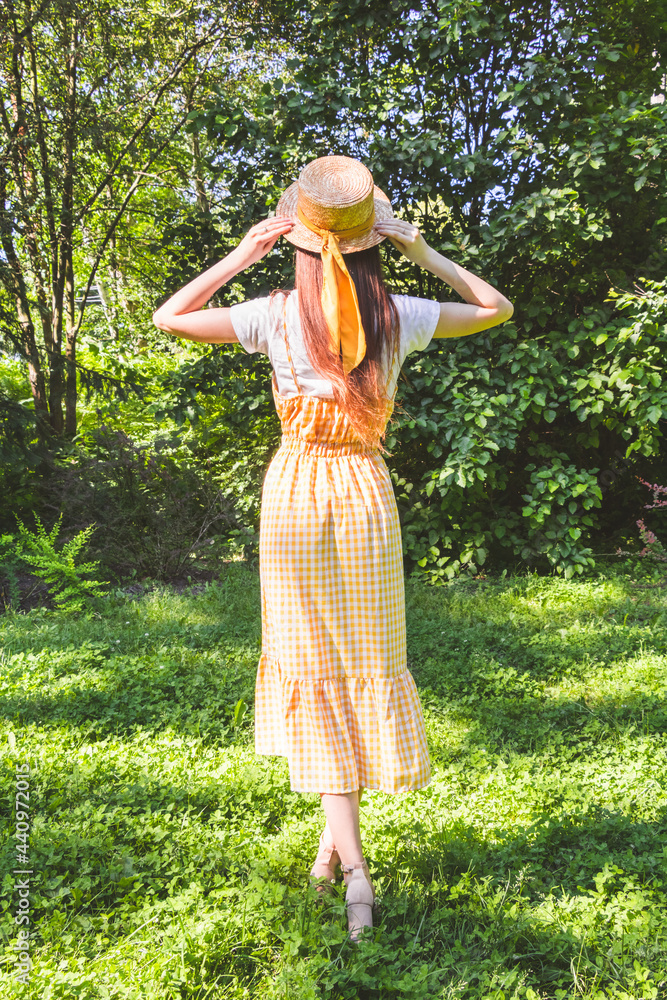 A girl in a hat with a ribbon and a yellow sundress with her back in full growth stands in a green garden