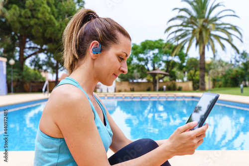 Young fitness woman choosing an online sport class outdoors. Horizontal side view of young woman connecting with technology to a gym class with friends next to a swimming pool.