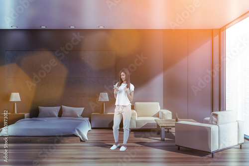 Vászonkép Attractive woman in cozy home wear browesing internet using smart phone in modern panoramic studio apartment with bed and living areas