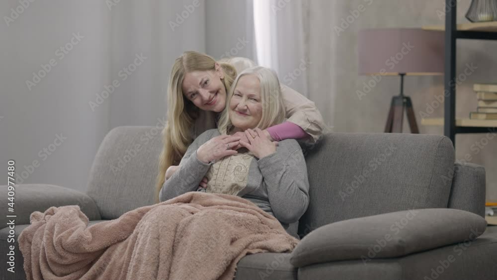 Wide shot portrait of smiling happy old woman sitting on couch as senior lady hugging mother looking at camera. Confident relaxed Caucasian retiree enjoying weekend with daughter at home posing Wide shot portrait of smiling happy old woman sitting on couch as senior lady hugging mother looking at camera. Confident relaxed Caucasian retiree enjoying weekend with daughter at home posing