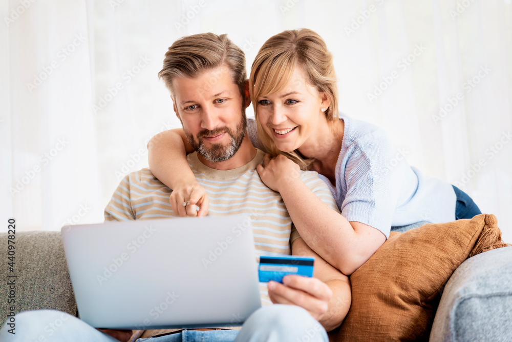Happy couple sitting on the couch at home while using laptop and bank card
