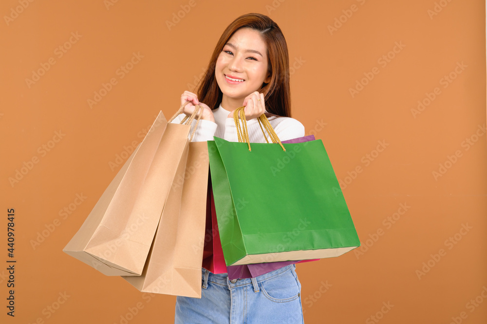 Beautiful young Asian woman wearing a white silk blouse, jeans, holding a shopping bag, smiling with a good mood, cheerful and cheerful in Holiday at the mall Center. With a brown background.
