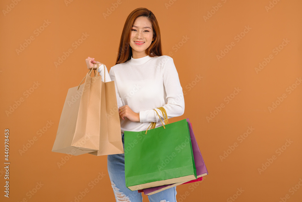 Beautiful young Asian woman wearing a white silk blouse, jeans, holding a shopping bag, smiling with a good mood, cheerful and cheerful in Holiday at the mall Center. With a brown background.