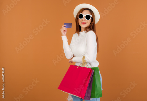Beautiful young Asian woman wearing white silk blouse and jeans, holding a shopping bag, Wear glasses holding credit card, smile good mood, Shopping in Holiday at shop in market. brown background. © Greanlnw studio