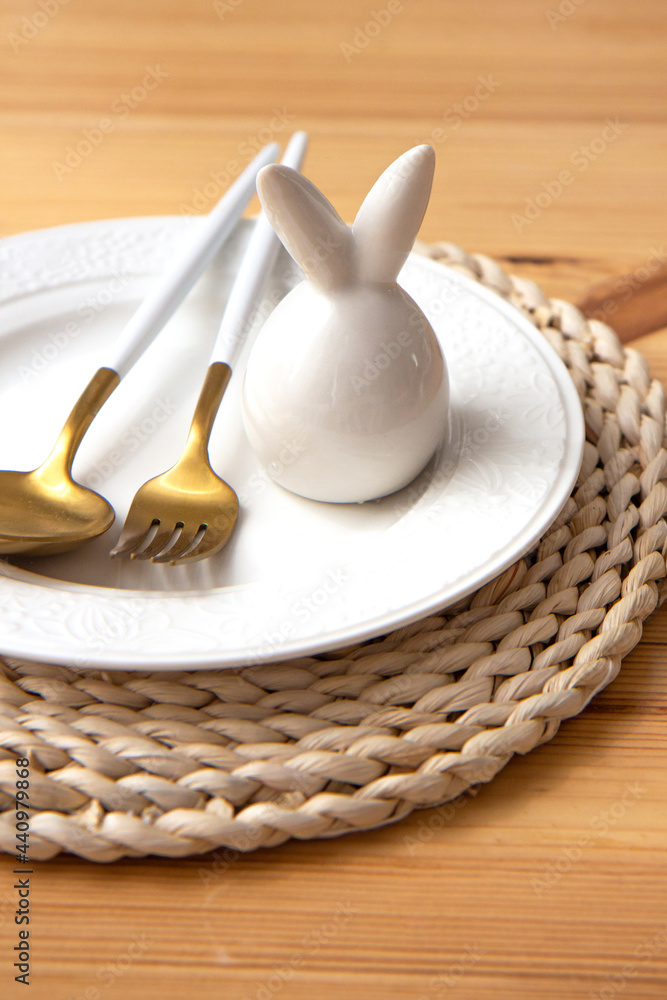 Cutlery golden spoon and fork and ceramic easter bunny lie on a white plate on an eco stand on a wooden table
