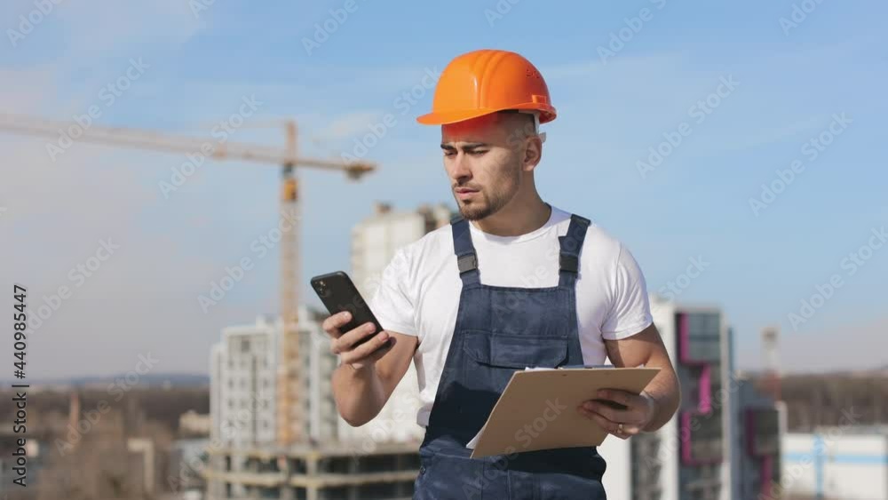 The young engineer is texting on his smartphone. He is focused and holding a folder of documents. He is wearing work clothes and a hard hat. He is on the roof of a business center. Construction in the