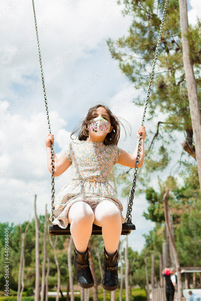 Little child playing on the swings in the park. She is wearing a face mask. Space for text.