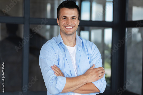 Startup concept, successful young caucasian businessman with arms crossed standing in the office, smiling, looks at the camera