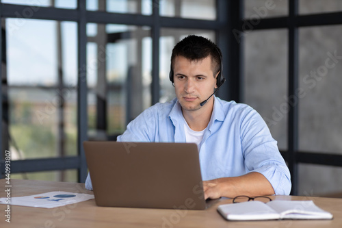 Concentrated male student with a headset looking at laptop screen sitting at the office table, online education, support worker, hot line