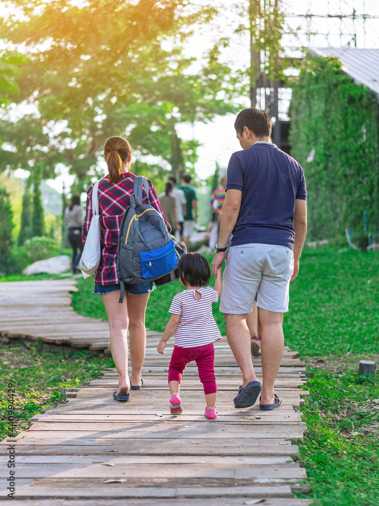 Fototapeta premium Back view of Asian Father hand holding lovely daughter walking on pathway through green garden.Dad and daughter walking together in park. Happy family spending time together outside in green nature.