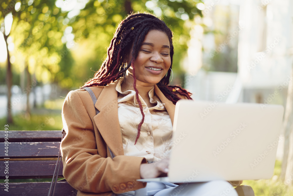 Fototapeta premium African american woman freelancer sitting on a bench in a street and using laptop