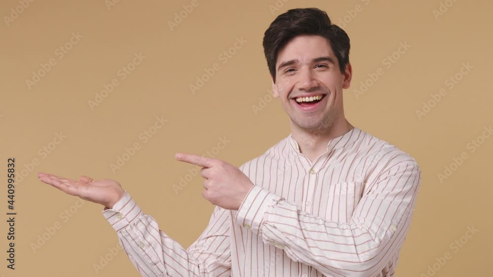 Stylish young promoter man 20s years old wears white striped shirt pointing fingers hands aside on workspace copy space mockup promo commercial area isolated on pastel beige background studio portrait Stylish young promoter man 20s years old wears white striped shirt pointing fingers hands aside on workspace copy space mockup promo commercial area isolated on pastel beige background studio portrait