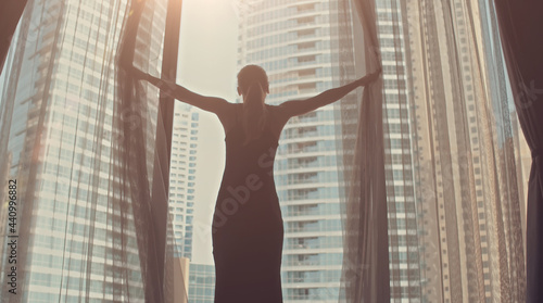 Canvas Print Silhouette of young woman opens curtains on the big window stretches arms and looking out her apartment on the city buildings during amazing sunrise