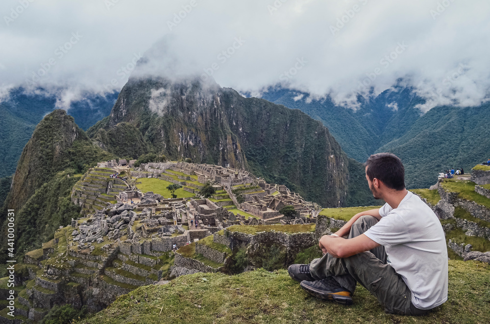Young man sitting, contemplating Machu Picchu lost city with Huayna Picchu mountain. Ruins of ancient inca civilization in the sacred valley of Cusco Province. Peru, South America Young man sitting, contemplating Machu Picchu lost city with Huayna Picchu mountain. Ruins of ancient inca civilization in the sacred valley of Cusco Province. Peru, South America