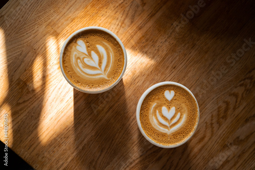 Two cups of fresh coffee with latte art on wooden table in sunlight. Morning cappuccino with hearts in cafe for couple. Coffee break concept. Caffeine drink for breakfast closeup. Top view, flat lay. 